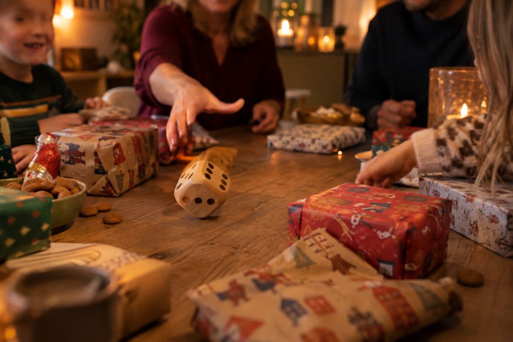 Gezin speelt een dobbelspel rond een tafel vol pakjes, een grote houten dobbelsteen rolt